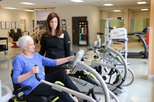 Female patient works on endurance, mobility and strength on the exercise bike in therapy gym with guidance from her female physical therapist