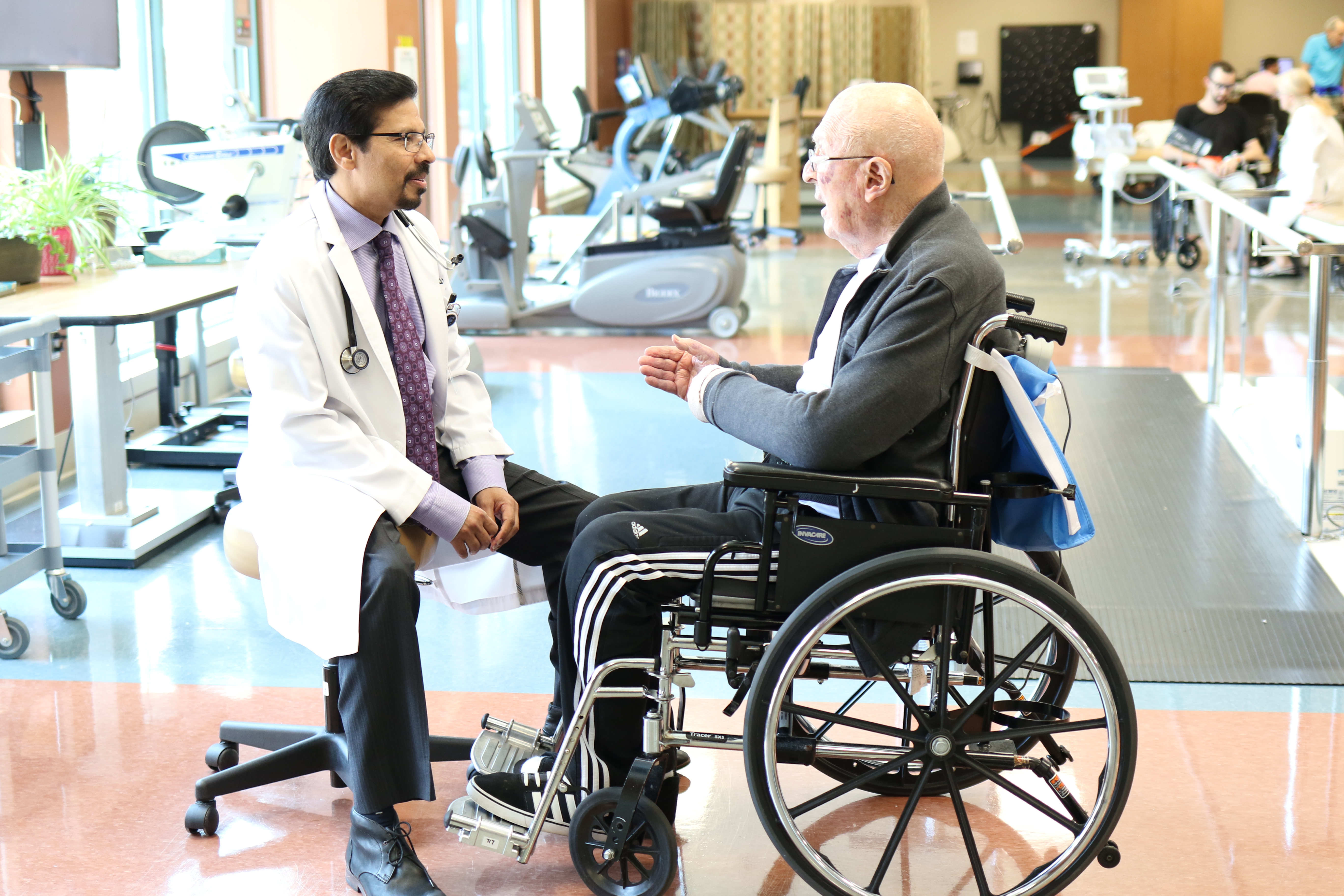 Male physician is seated and communicating with a male patient seated in a wheelchair. The conversation is taking place in the therapy gym where other therapies are taking place.