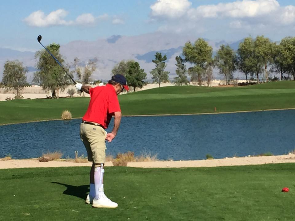 Man golfing on the greens near water in a red shirt, black hat and khaki shorts