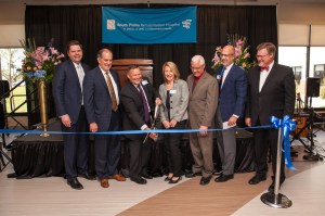 Seven people in business attire look on as on man cuts blue ribbon with large scissors