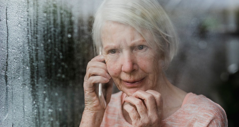 Senior woman is looking worriedly out of the window of her home while talking to someone on the phone.